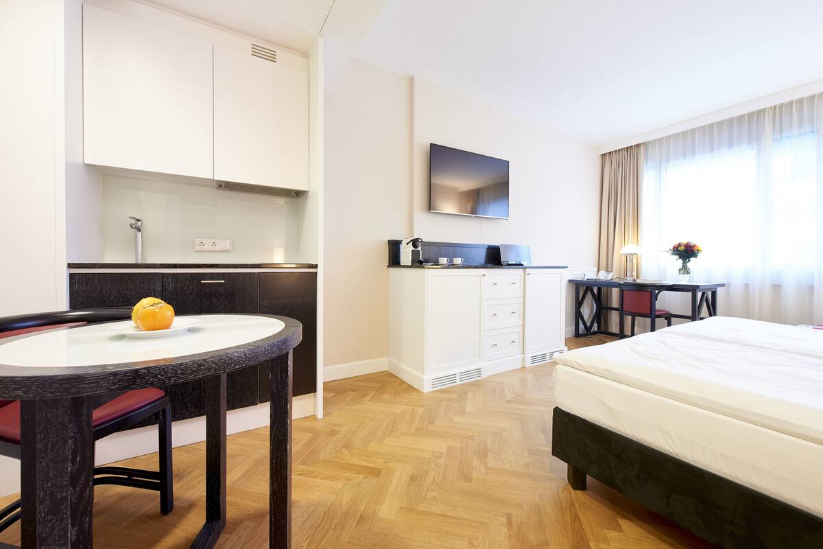 Ein Hotelzimmer mit Bett, Schreibtisch mit Stuhl, einer kleinen Küchenzeile und einem runden Esstisch mit Stühlen.