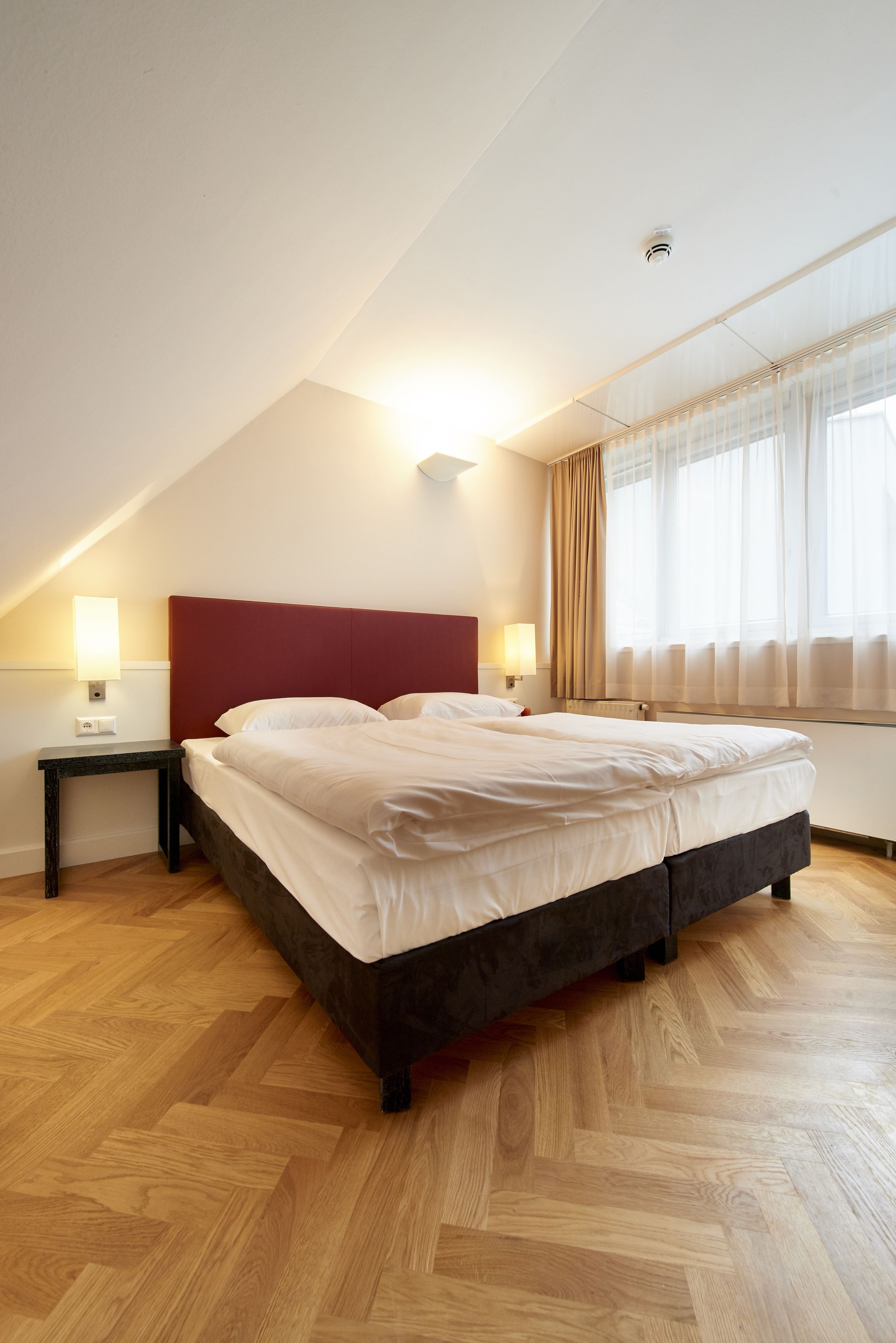 Un dormitorio con una cama grande, suelo de madera, cabecera roja y una ventana con cortinas.