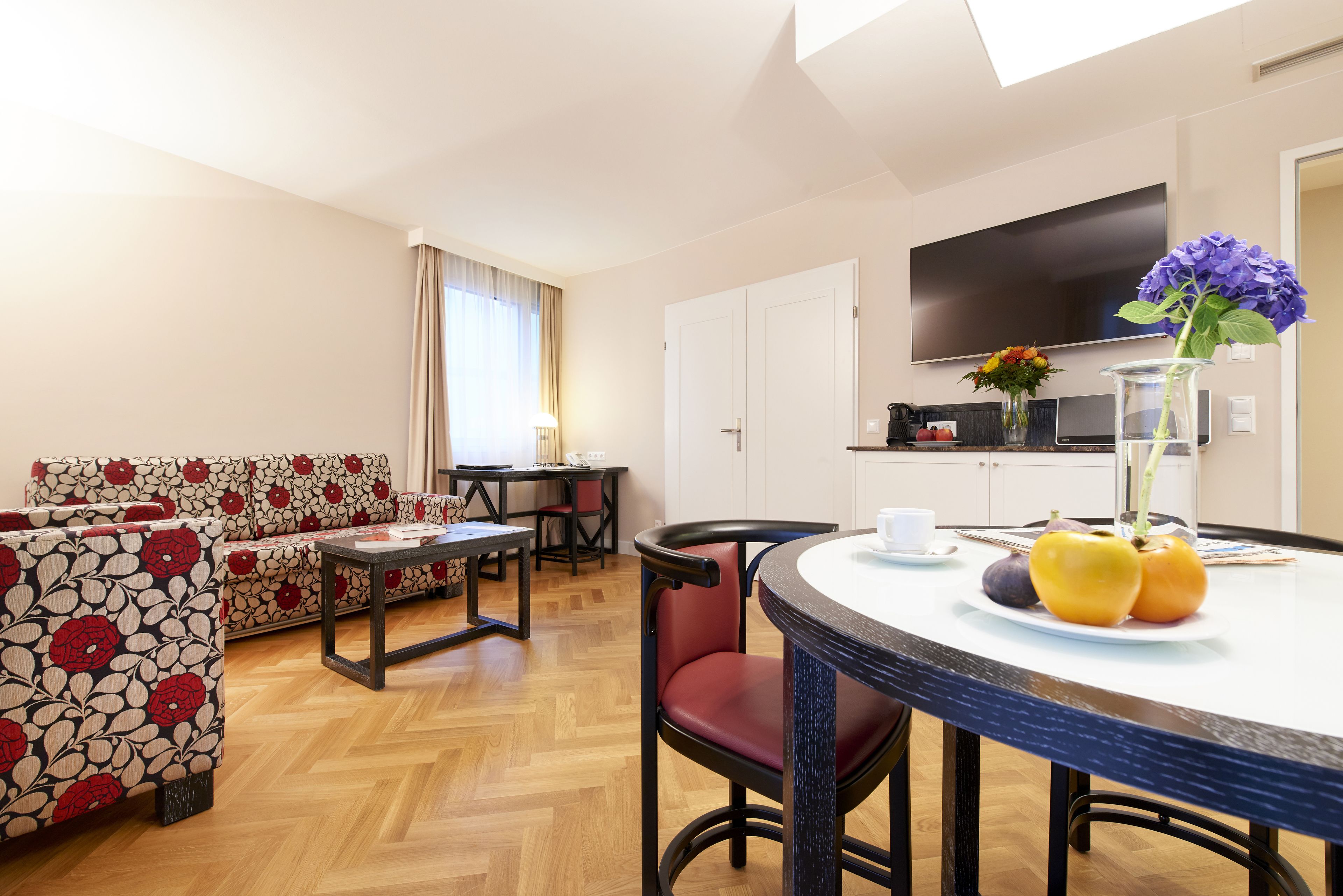 A modern living room with a sofa, dining table, and chairs. A flat-screen TV is mounted on the wall.