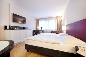 Quarto de hotel com cama, escrivaninha, cadeira, televisão e janela com cortinas.