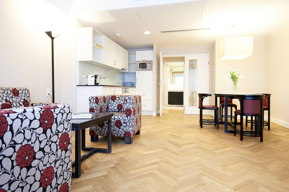 Ein modernes Wohnzimmer mit Fischgrätparkett, einem Essbereich mit gemusterten Sofas, einem Couchtisch und einer kleinen Küchenzeile mit weißen Schränken und einer Mikrowelle.