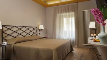 Habitación de hotel con una cama grande, una mesita de noche con una taza y platillo, una pequeña mesa con pasteles y un jarrón con flores.