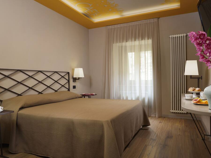 Habitación de hotel con una cama grande, una mesita de noche con una taza y platillo, una pequeña mesa con pasteles y un jarrón con flores.