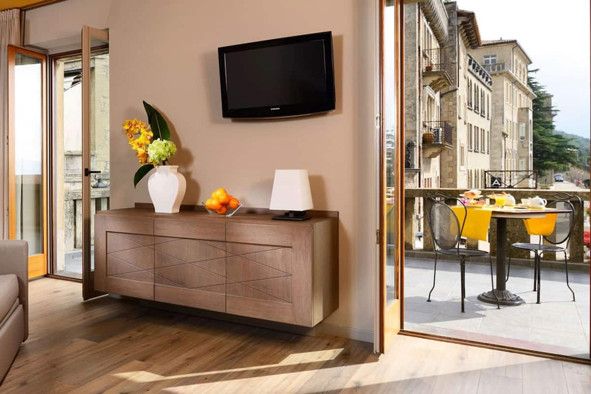 A modern hotel room with a sofa, a wooden console table with a vase and fruit, a TV, and a balcony with a table and chairs.
