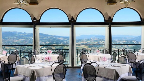 A restaurant with tables set for dining, large arched windows, and a view of the countryside. - Titano Suites (San Marino)
