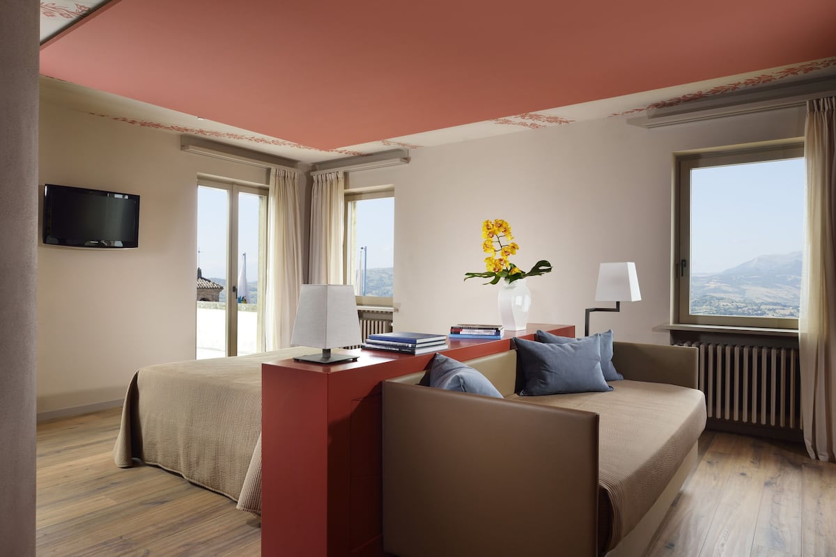 A hotel room with a bed, a sofa, a desk with a lamp, a TV, and a window with a view.