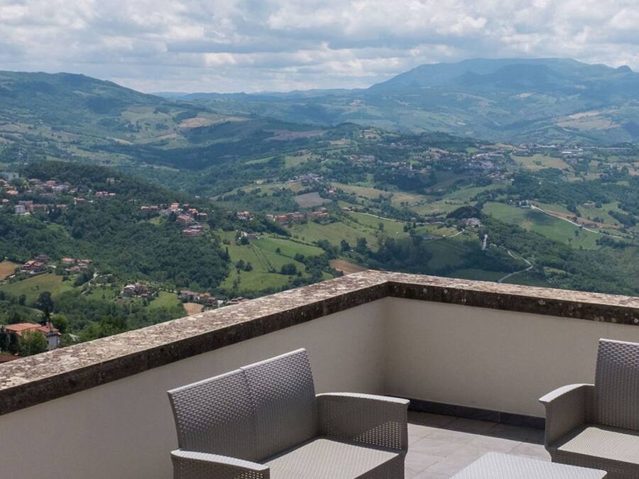 Ein Balkon mit Korbstühlen, der Blick auf ein Tal mit Häusern und Hügeln freigibt.