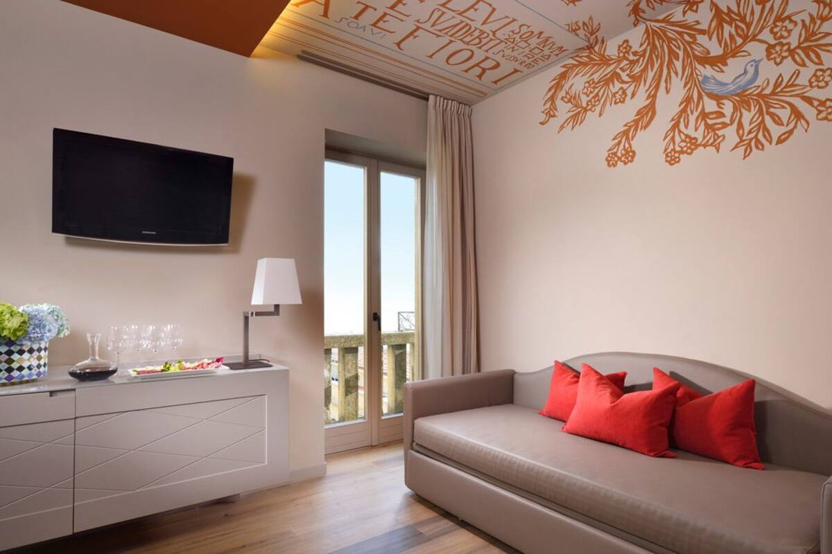 A modern living room with a flat-screen TV, a sofa with red cushions, a dining area with a vase and glasses, and a balcony with a view.