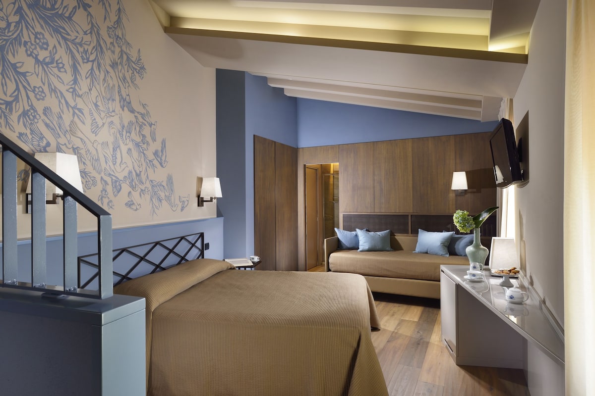 A hotel room with a large bed, a sofa, a desk with a flat-screen TV, and a wall-mounted TV.