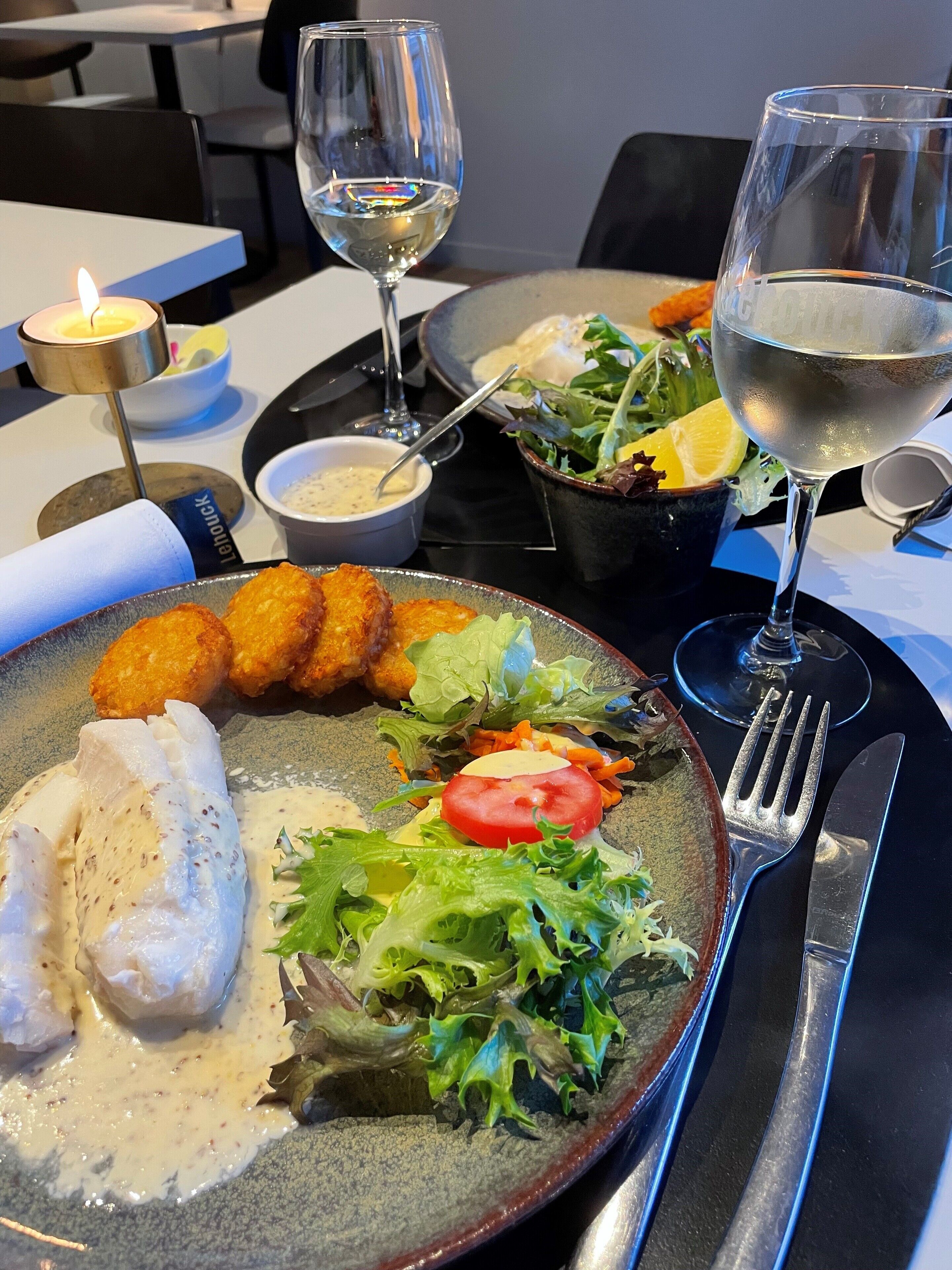 A dining table with a plate of food, including fish, salad, and bread rolls, and two glasses of white wine.