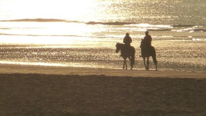 Duas pessoas a cavalo numa praia ao pôr do sol.