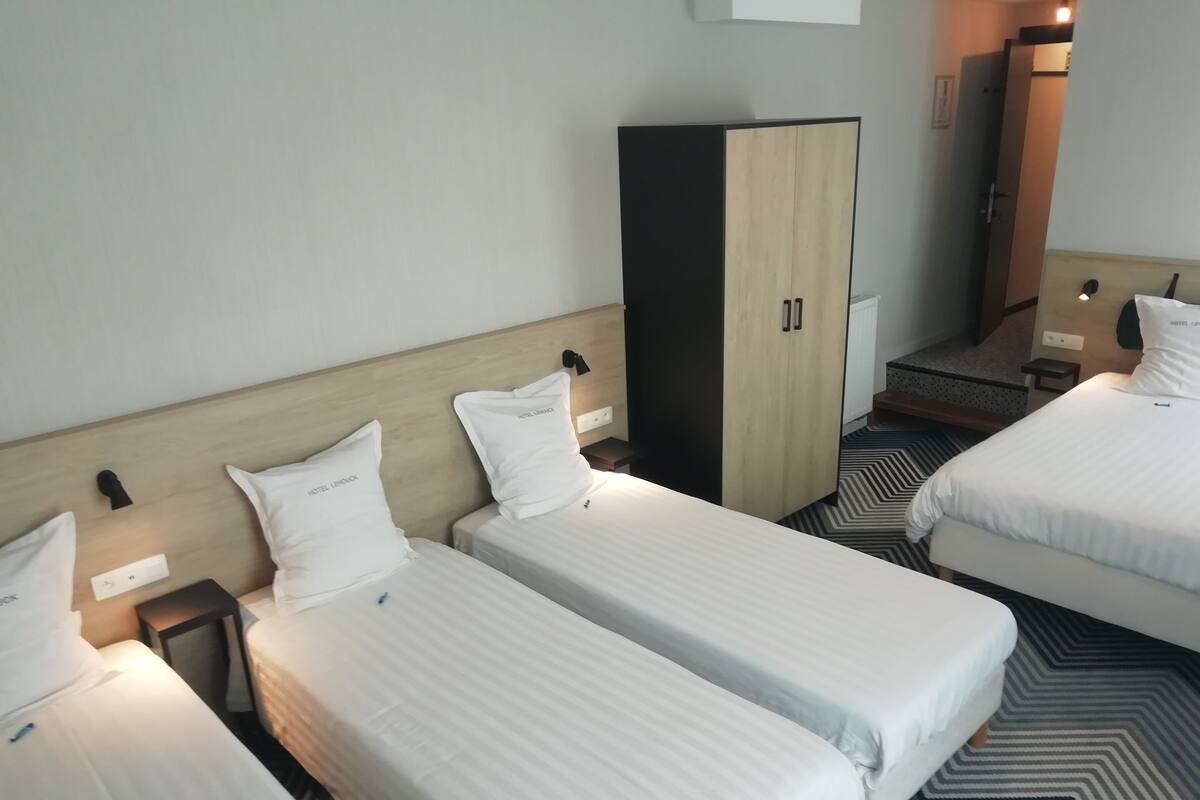 Ein Hotelzimmer mit drei Einzelbetten, jedes mit einem weißen Kopfkissen mit dem Aufdruck "HOTEL LONDON". Ein Kleiderschrank mit schwarzem Rahmen und heller Holzoptik sowie ein gemustertes Teppichboden sind ebenfalls vorhanden.