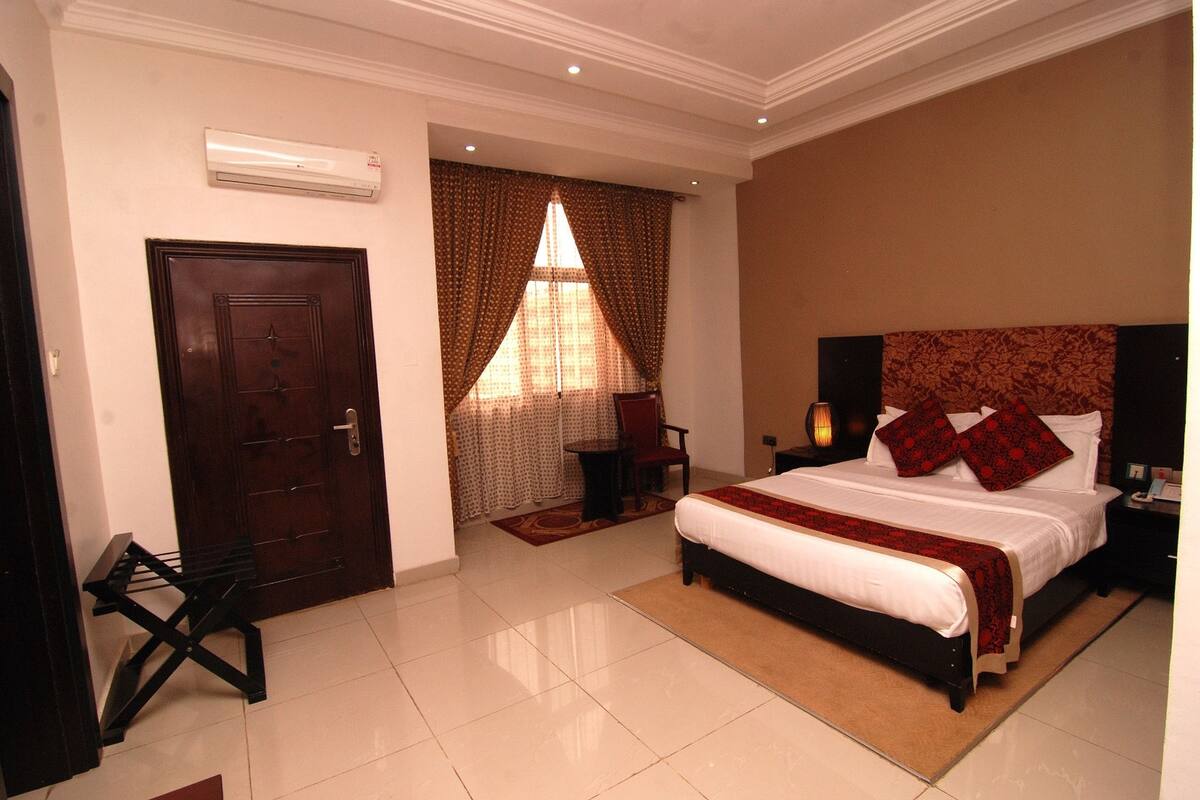 A hotel room with a large bed, a wooden door, a small table, and a window with curtains.