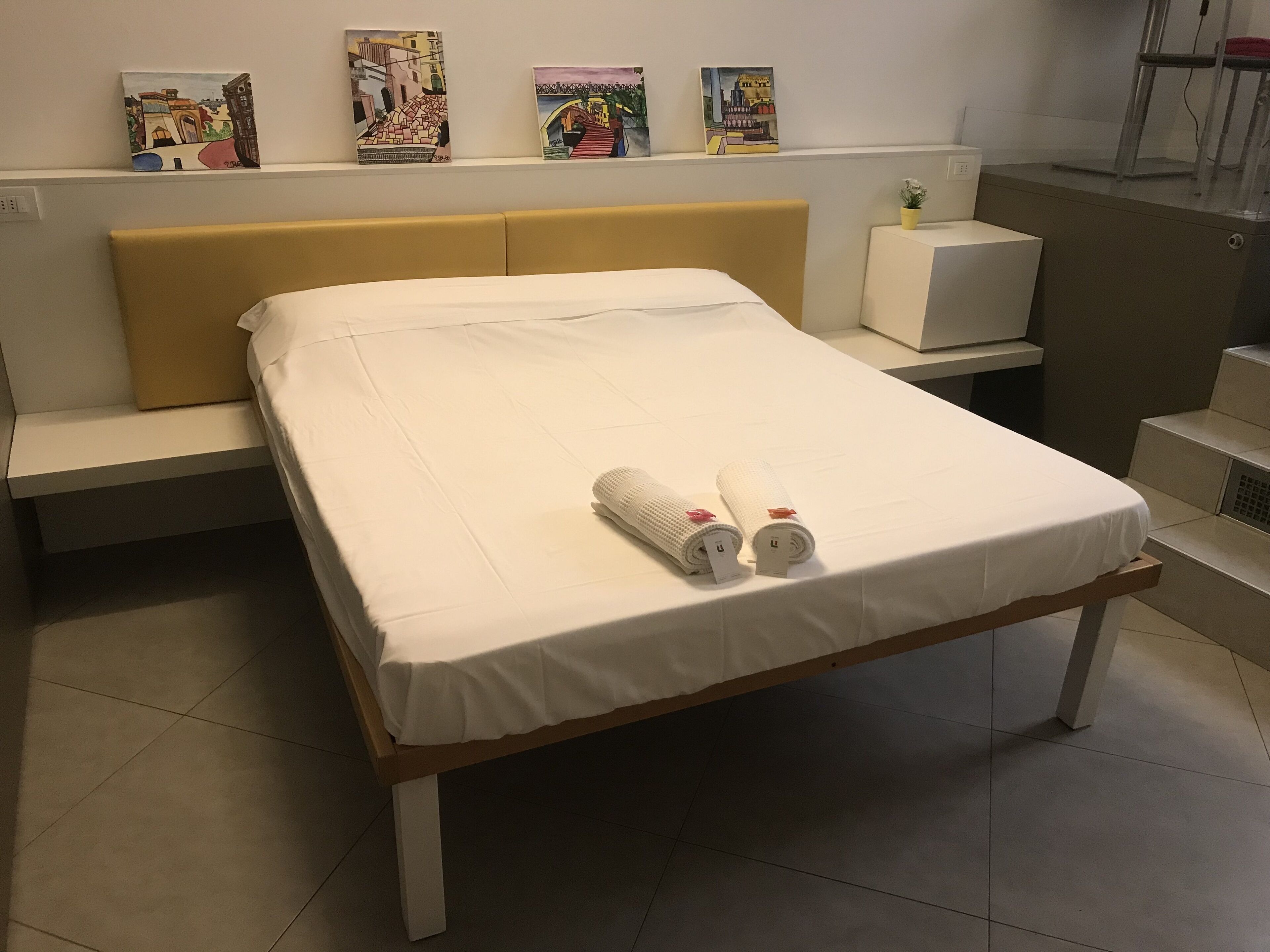 Una cama bien tendida con sábanas blancas y dos toallas enrolladas, un cabecero amarillo y una mesita de noche con una planta en maceta.