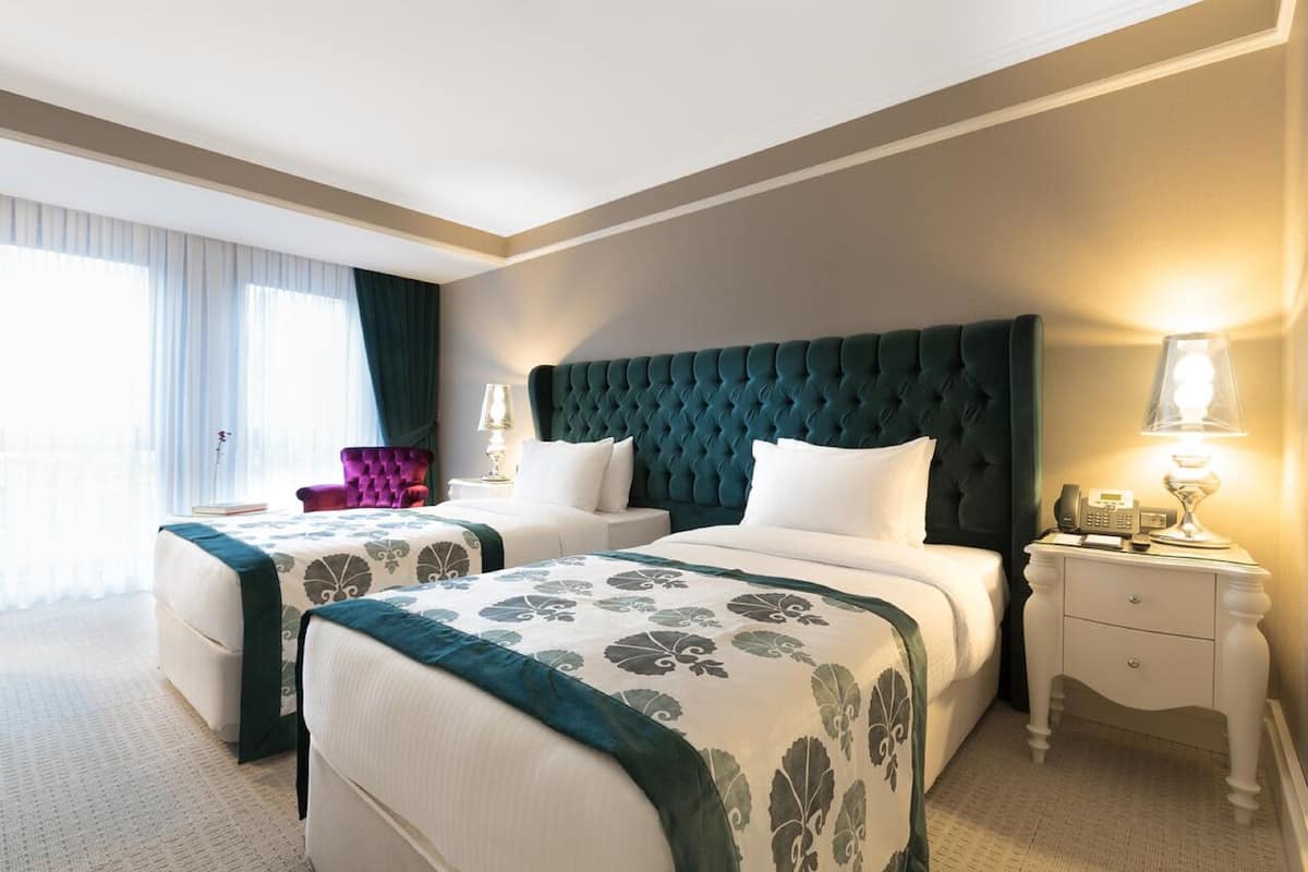 A hotel room with two beds, a nightstand with a lamp, a chair, and a large window with curtains.