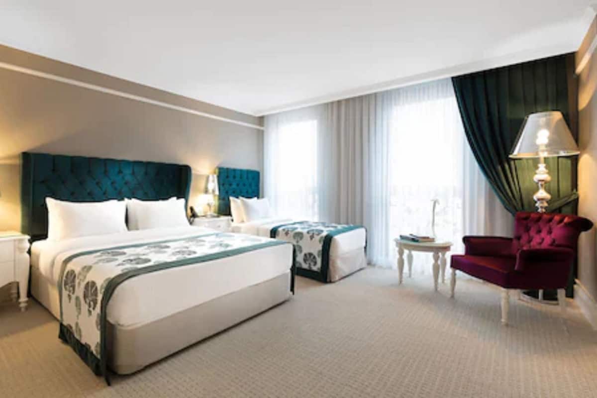 A hotel room with two beds, a red armchair, a small table, and a lamp.