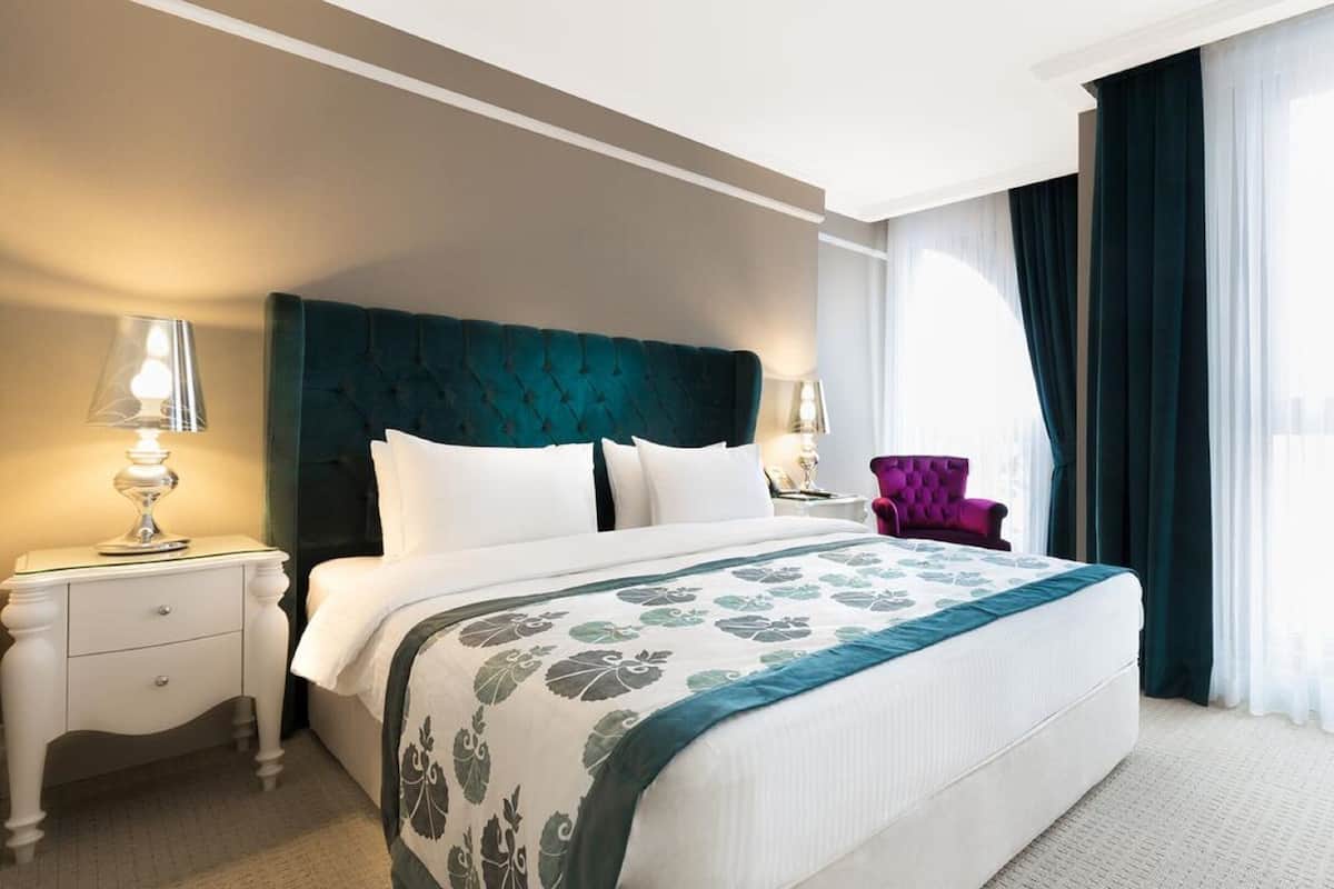 A hotel room with a large bed, two bedside tables, a nightstand, a purple armchair, and a window with curtains.