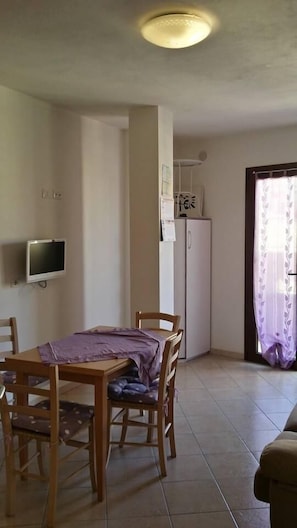 A dining area with a wooden table and chairs, a refrigerator, a television mounted on the wall, and a door with a curtain.
