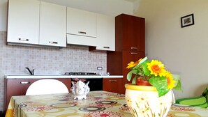 Una cocina con gabinetes blancos, salpicadero de azulejos, estufa y pileta. Hay una mesa con mantel floral y un florero con flores.
