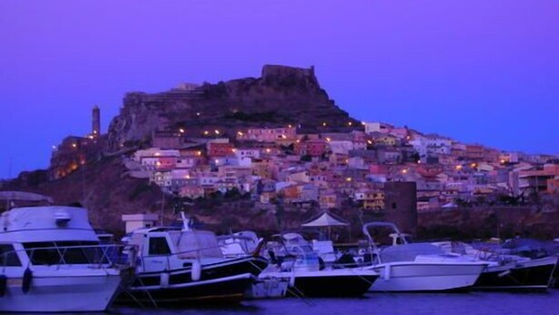 Un puerto con barcos amarrados y un asentamiento en la ladera, muy poblado, al fondo.