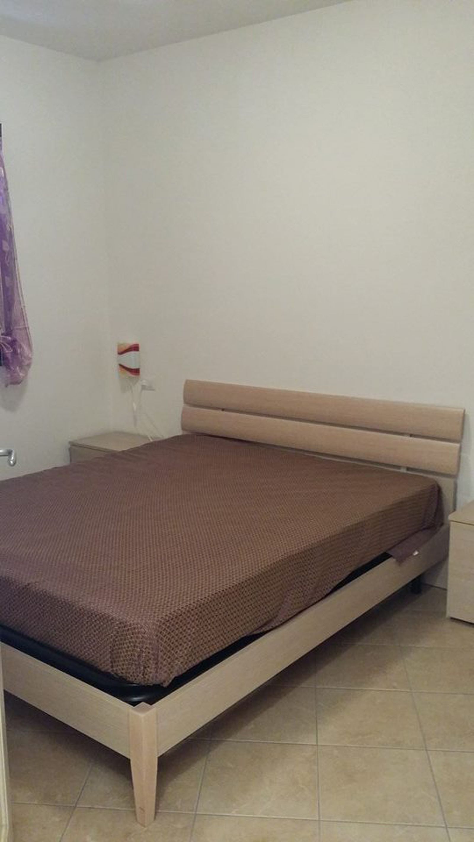 Una cama individual con estructura de madera y colchón marrón en una habitación con paredes blancas y suelo de baldosas.