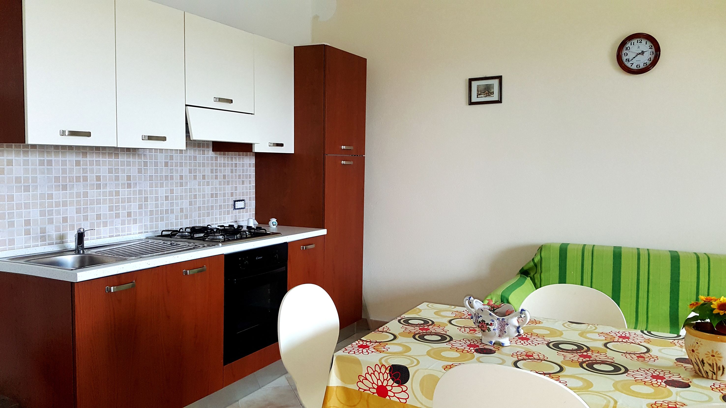 A compact kitchen with white cabinets, a gas stove, and a dishwasher. There's a dining table with chairs and a green-striped sofa. A clock and a framed picture adorn the wall.