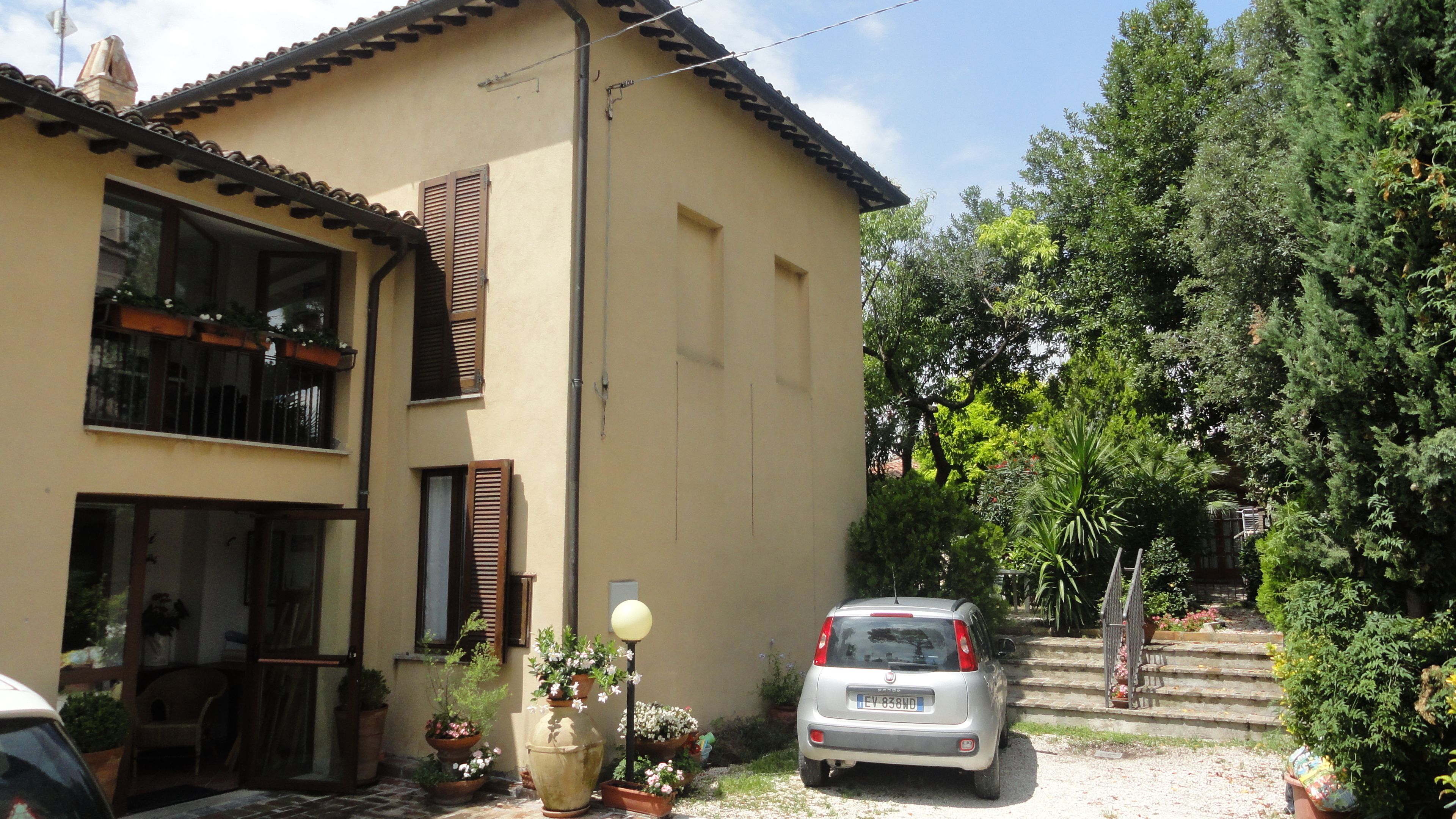 A two-story house with a car parked in the driveway, surrounded by potted plants and trees.