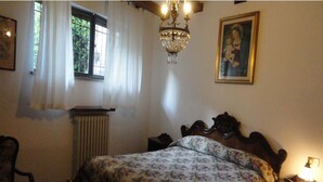 A bedroom with a wooden bed, a chandelier, a window with curtains, a radiator, and a framed picture on the wall. - La Terrazza di spello (Spello)