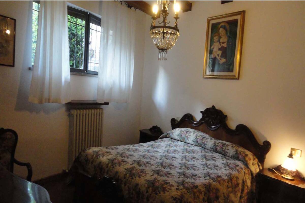 A bedroom with a wooden bed, a chandelier, a window with curtains, a radiator, and a framed picture on the wall.