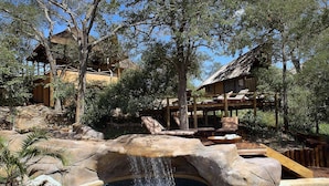 A pool with a waterfall, surrounded by large rocks and trees, with wooden structures and lounge chairs in the background. - Lukimbi Safari Lodge (Nkomazi)