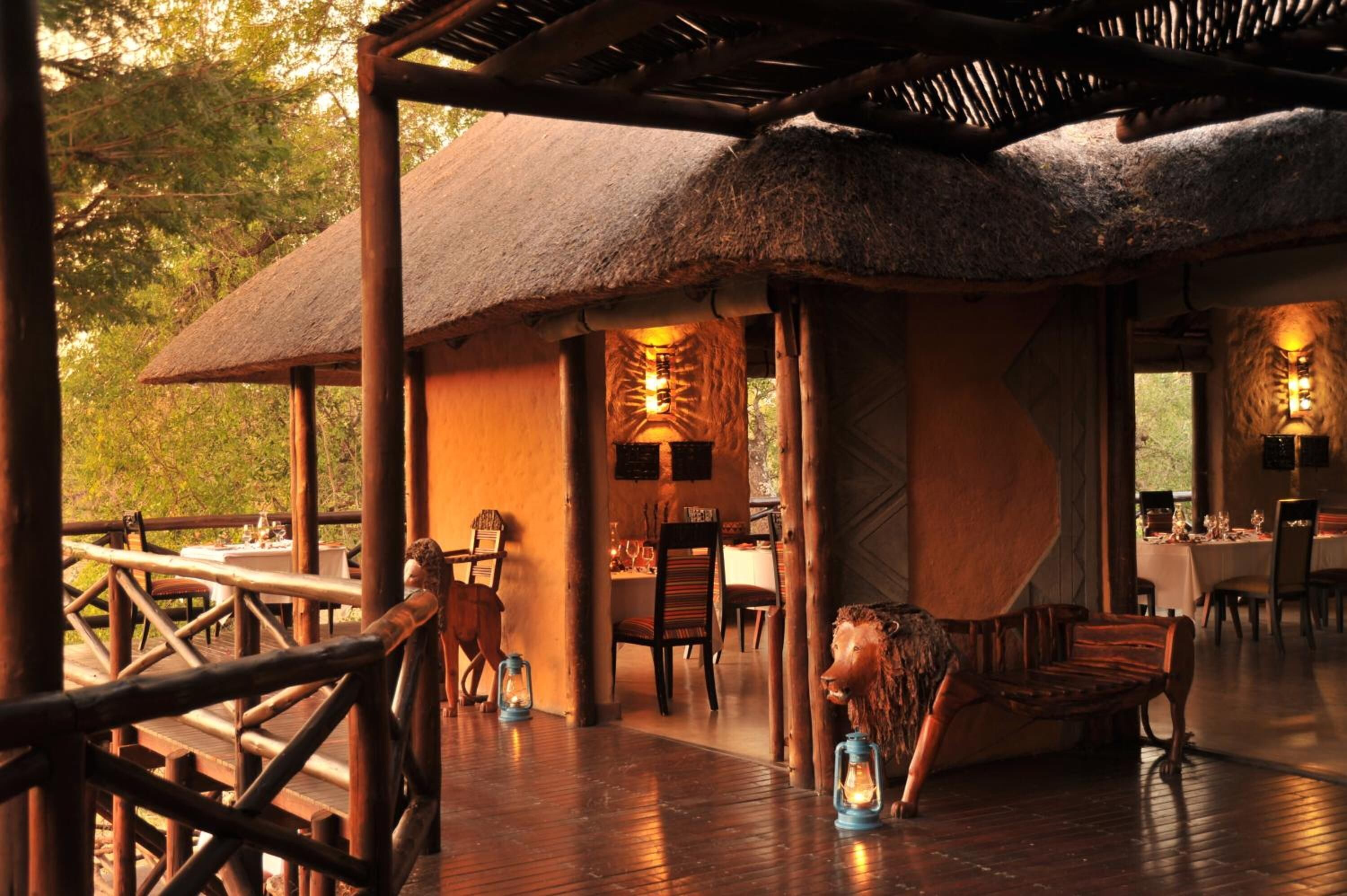 A thatched-roof dining area with wooden furniture and lanterns, surrounded by a wooden deck and natural scenery.
