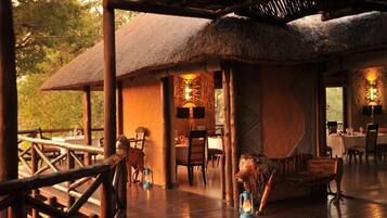 A thatched-roof dining area with wooden furniture and lanterns, surrounded by a wooden deck and natural scenery.