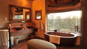 A bathroom with a large bathtub, two sinks, a large mirror, and a window offering a view of trees. - Lukimbi Safari Lodge (Nkomazi)