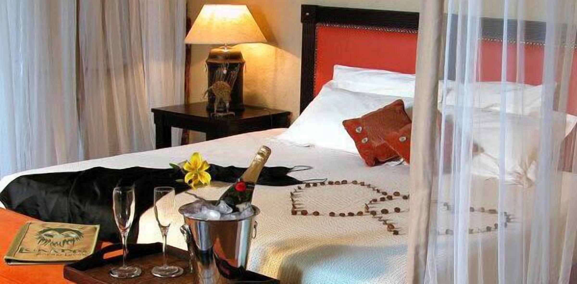 A hotel room with a bed, a nightstand with a lamp, a bottle of champagne, and a flower.