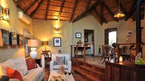 A cozy living room with a high ceiling, a sofa, a coffee table with a tray of fruits and wine glasses, and a wooden dining area with chairs. - Lukimbi Safari Lodge (Nkomazi)