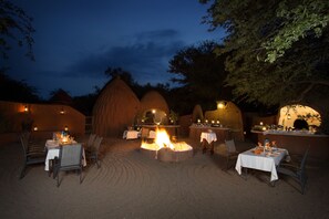 An outdoor dining area with round tables and chairs, a central fire feature, and a rustic architectural backdrop.