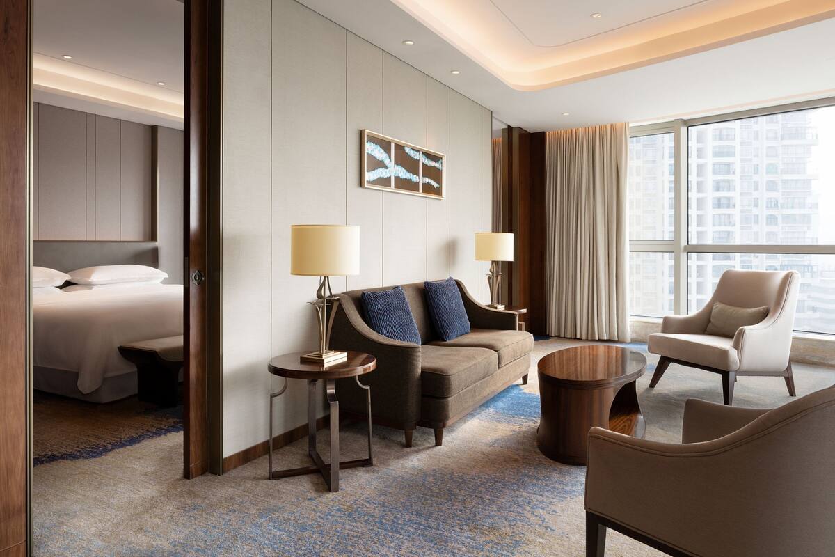 Una habitación de hotel moderna con cama, sofá, sillón, dos mesitas auxiliares y un ventanal con vistas a la ciudad.