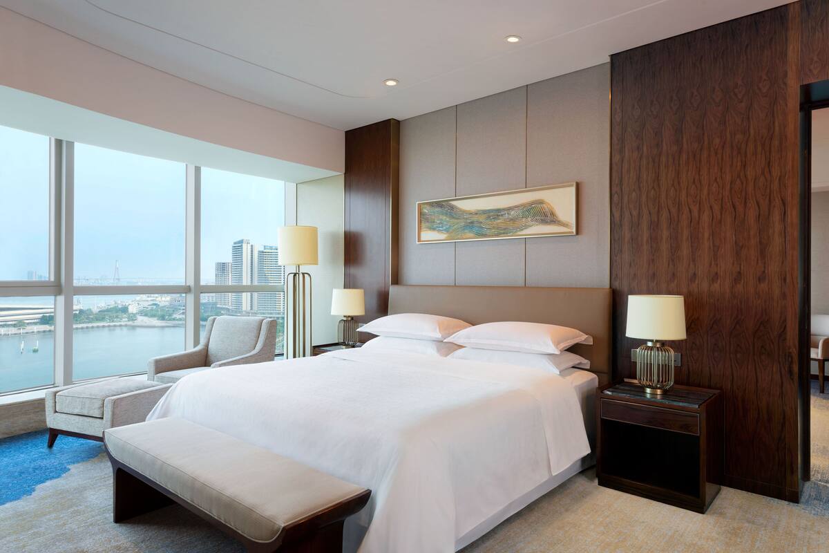 Una habitación de hotel moderna con una cama grande, dos mesitas de noche con lámparas, una silla y vistas a la ciudad a través de amplias ventanas.