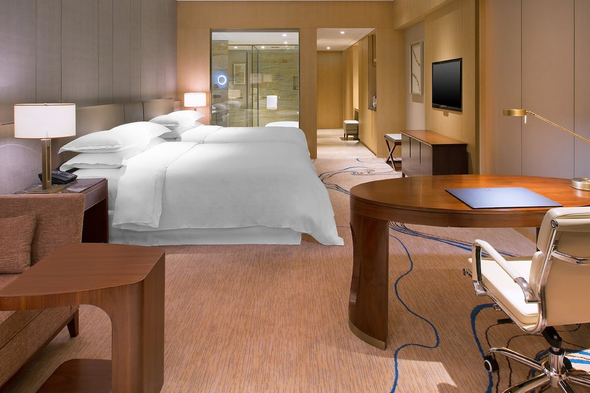 Habitación de hotel con una cama grande, un escritorio, una silla, un televisor y un baño visible a través de una puerta de cristal.