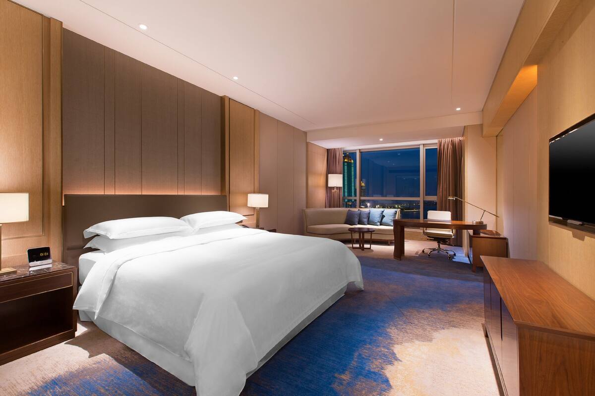 Habitación de hotel con una cama grande, un escritorio, una silla, un televisor y vistas de la ciudad de noche.