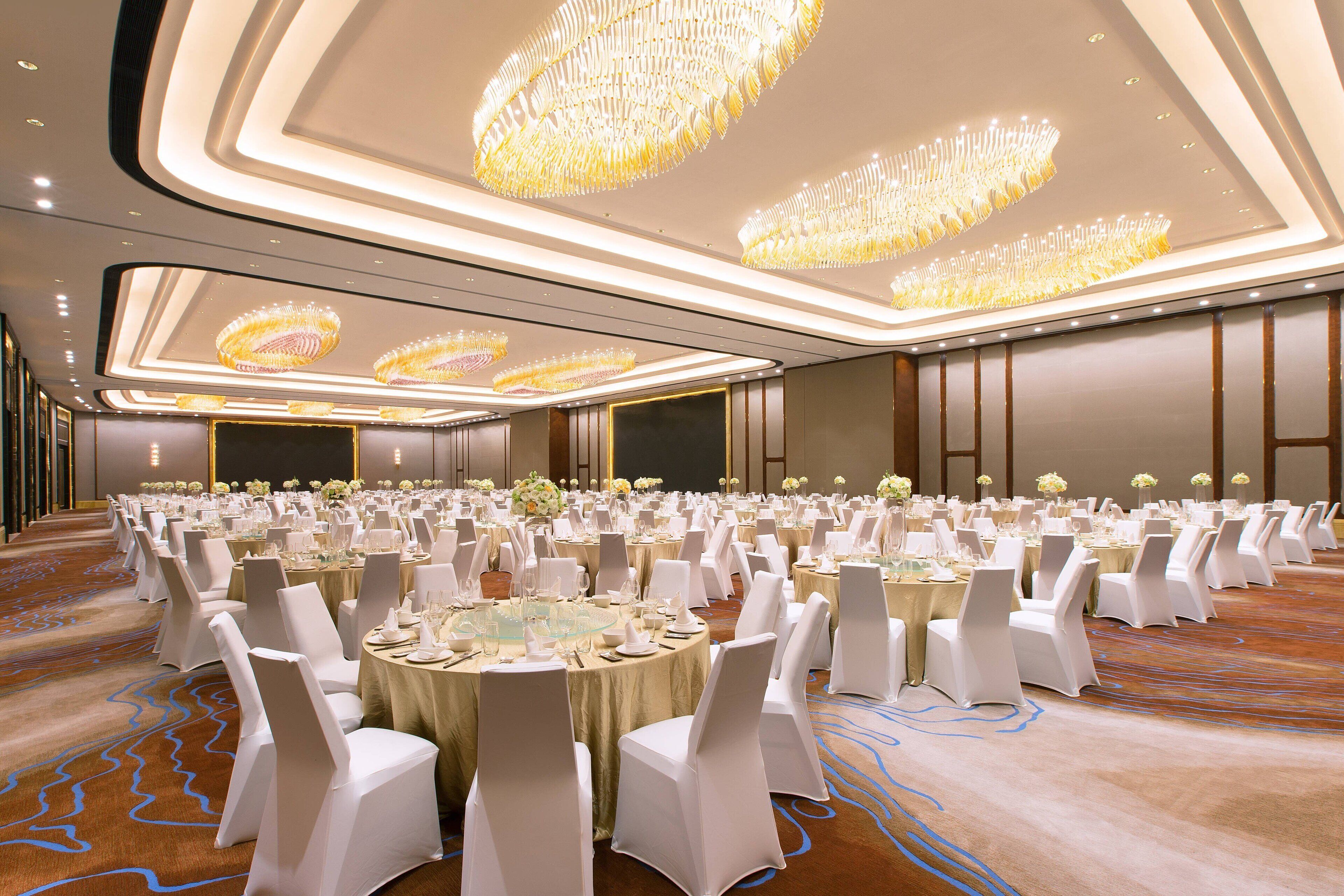 A banquet hall with round tables, white chairs, and chandeliers.