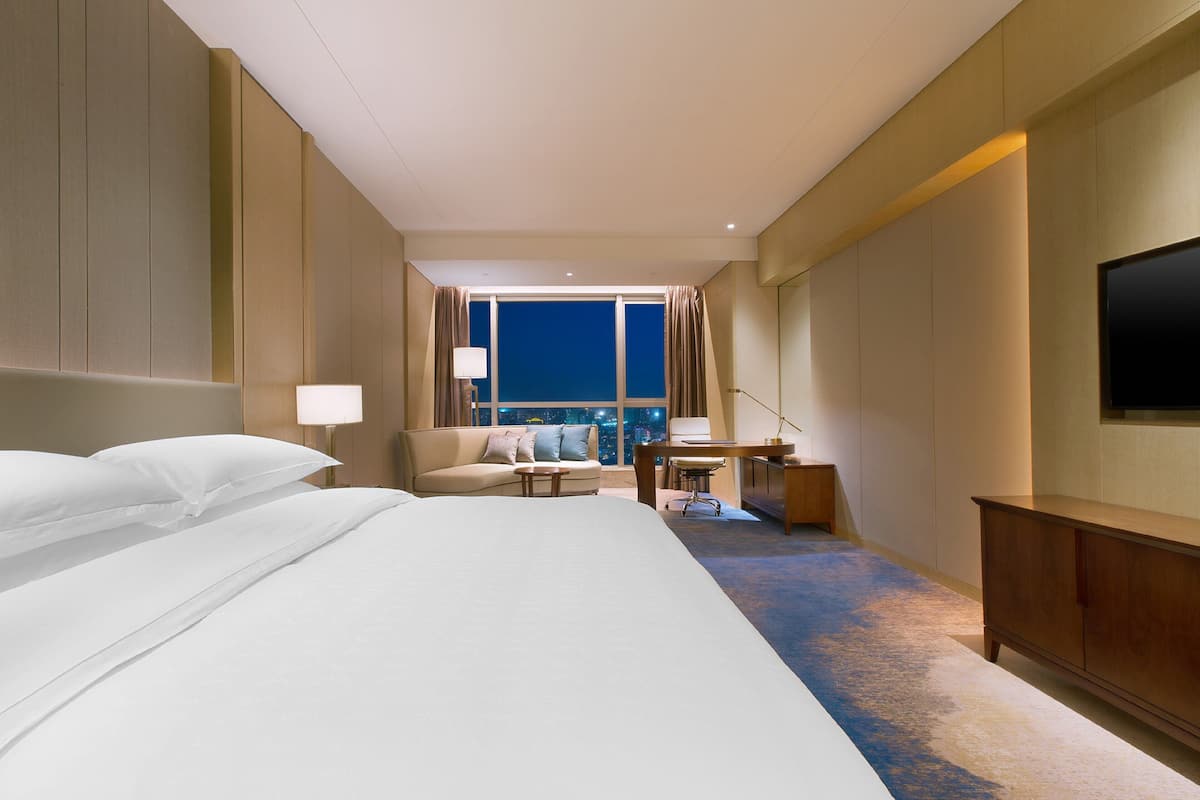 Habitación de hotel con una cama grande, un escritorio, una silla, un televisor y vistas a la ciudad de noche.
