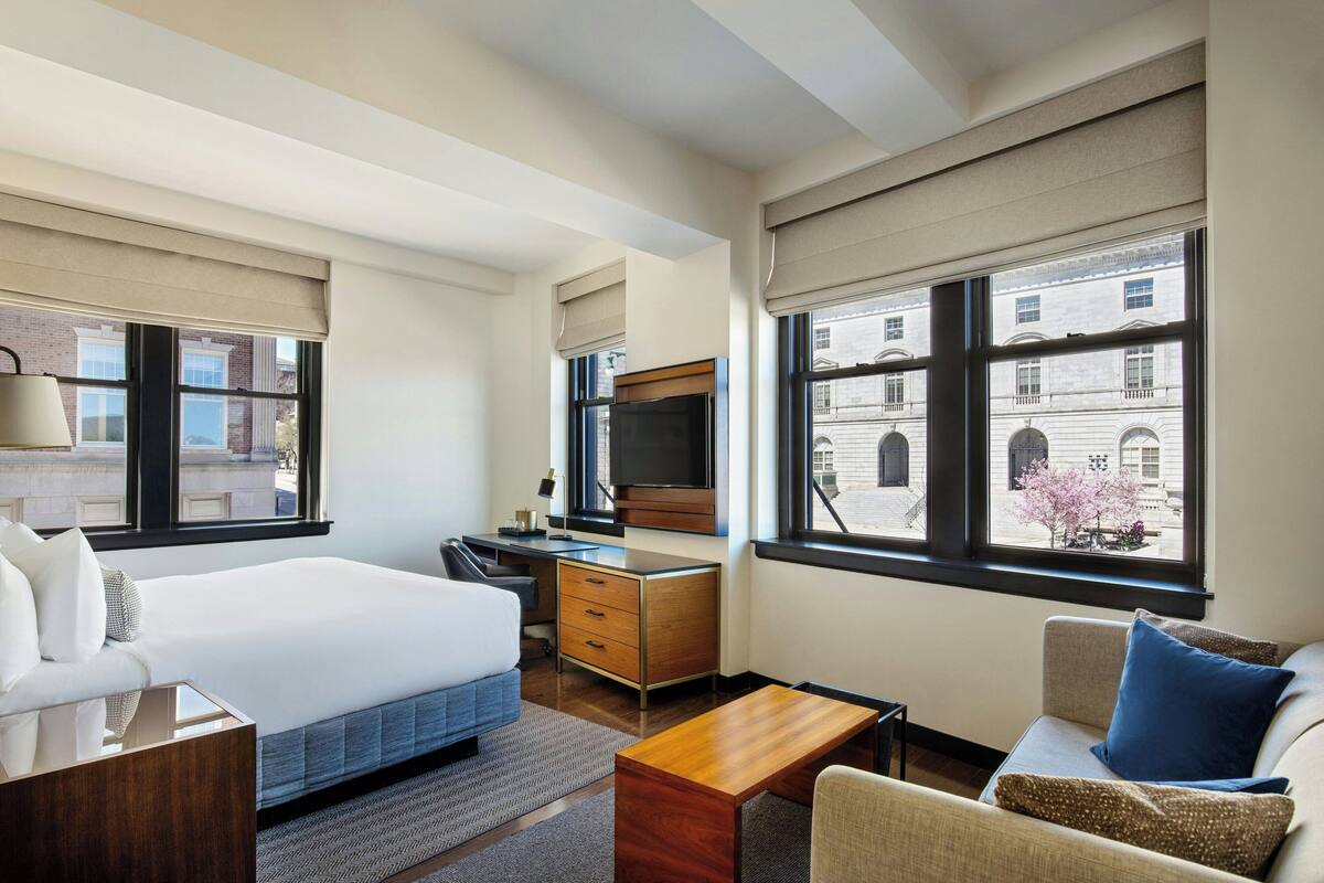 Quarto de hotel com cama, escrivaninha, televisão e vista para edifícios através de grandes janelas.