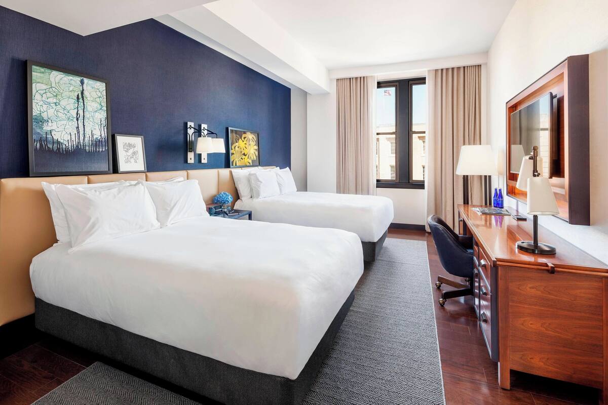 Quarto de hotel com duas camas, uma escrivaninha, uma cadeira e obras de arte nas paredes.