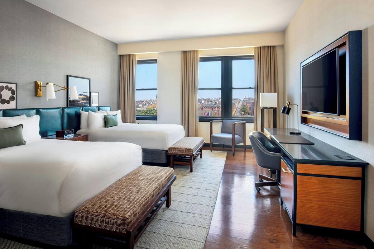 Quarto de hotel com duas camas, uma escrivaninha, uma cadeira, uma televisão e vista para a cidade.