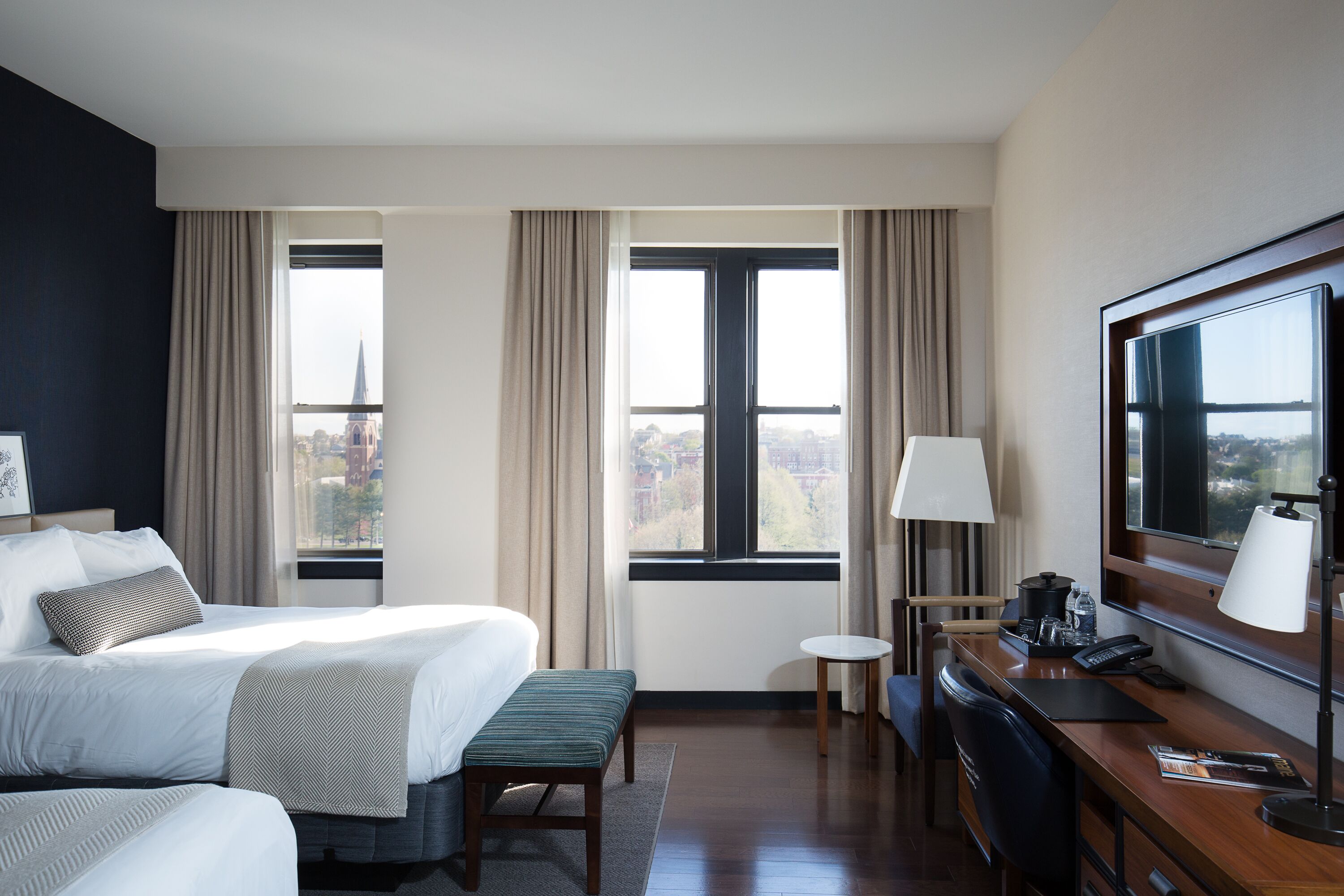 A hotel room with a large bed, a desk with a computer, a chair, a small table, and a large window with curtains.