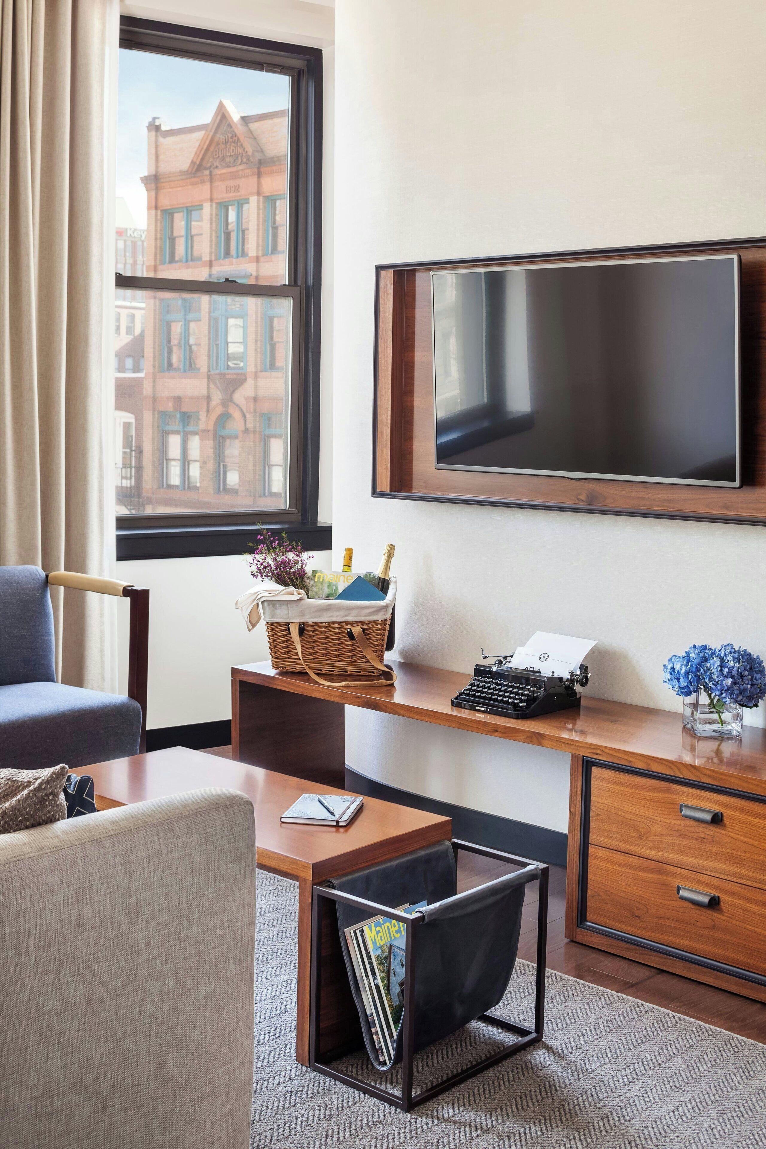 Quarto de hotel com uma janela grande, uma escrivaninha com uma máquina de escrever, uma cadeira, uma mesa pequena, uma televisão e uma estante de livros.