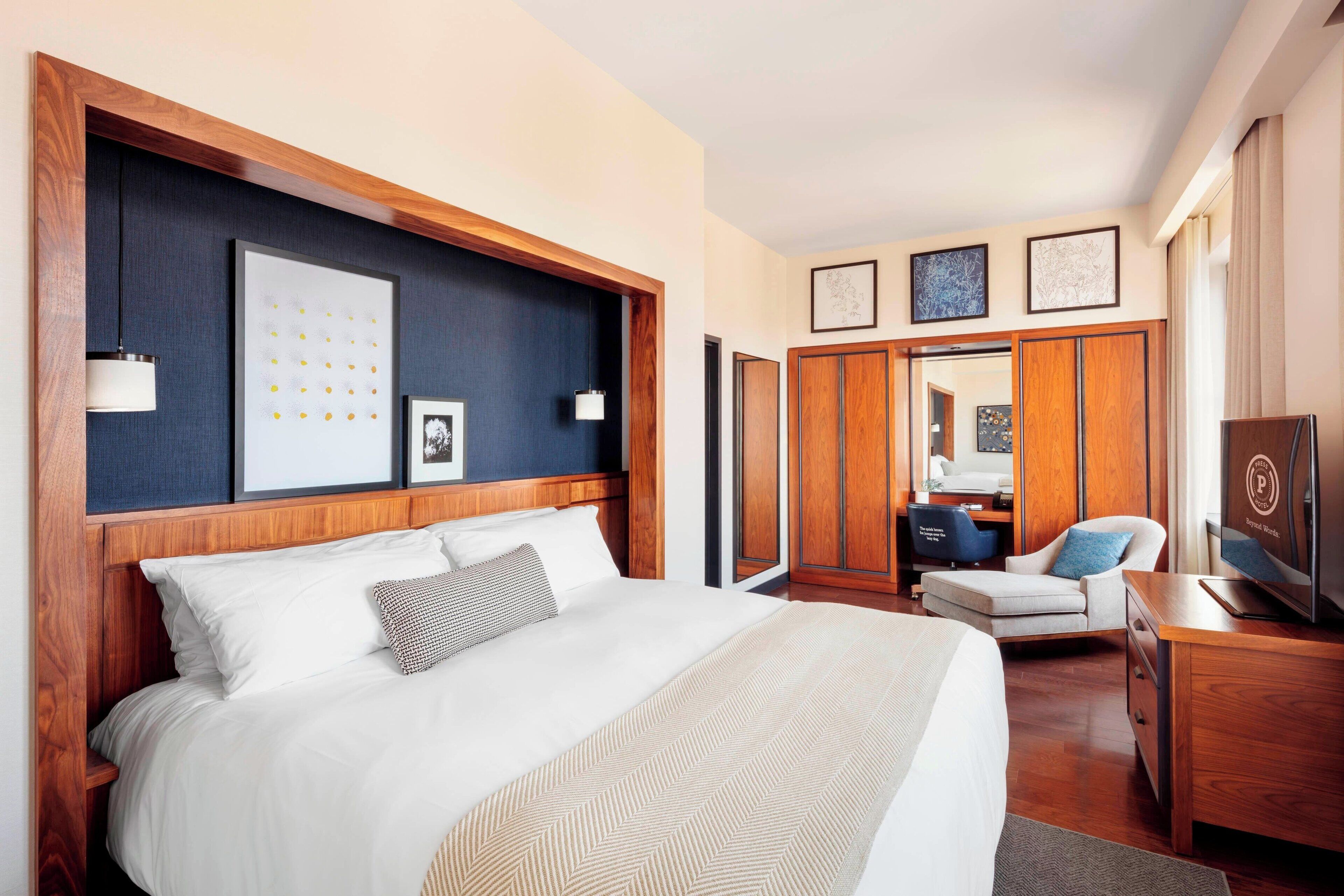 Une chambre d’hôtel avec un grand lit, une tête de lit en bois, une armoire, un coin salon avec un fauteuil et un téléviseur.