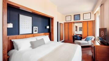 Une chambre d’hôtel avec un grand lit, une tête de lit en bois, une armoire, un coin salon avec un fauteuil et un téléviseur.