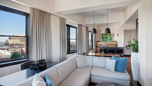 A modern living room with a large sectional sofa, a dining area with a kitchen in the background, and large windows offering a city view. - The Press Hotel, Marriott Autograph Collection (Portland)